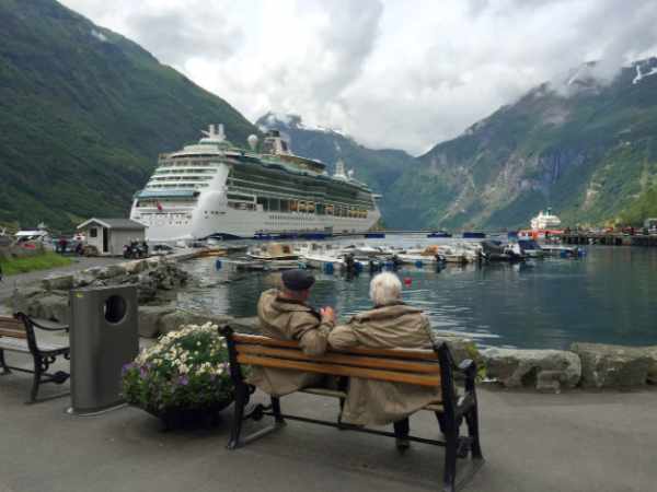 Photo of a retired couple sitting on a bench by a lake with a large cruise ship in the background